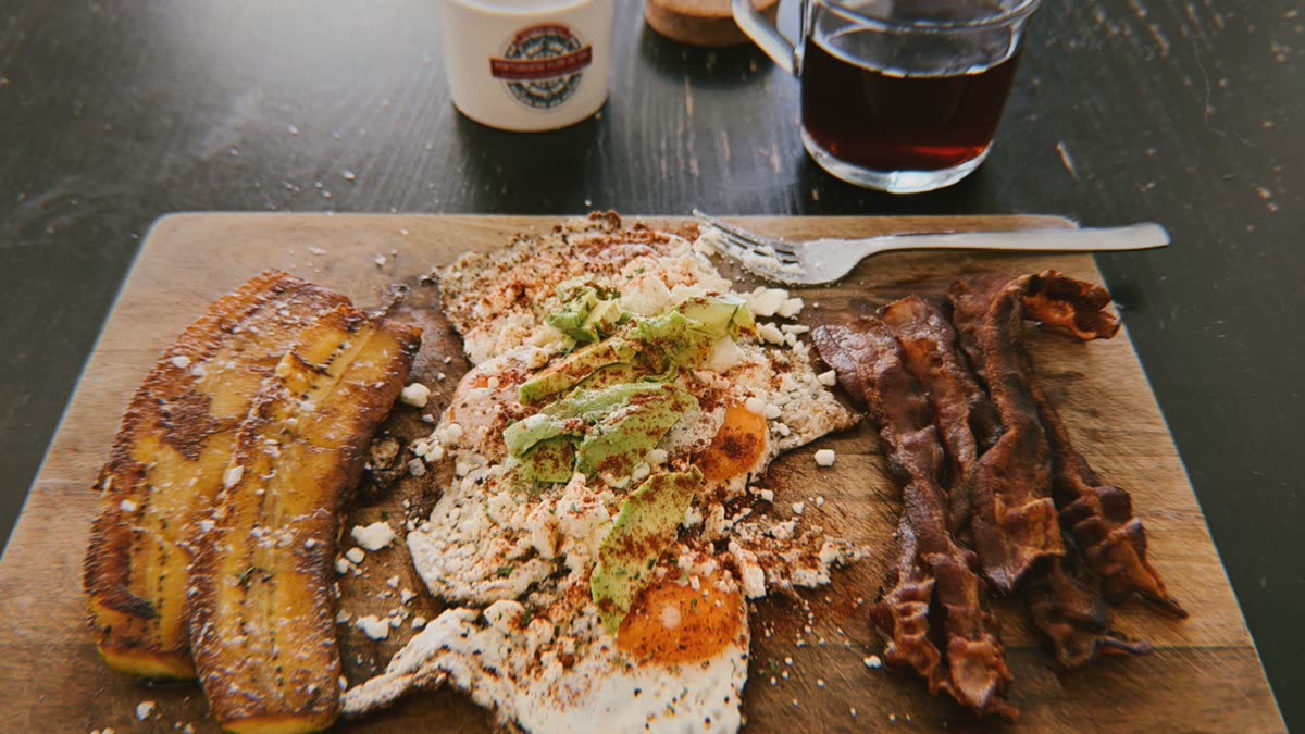 Plated primal lunch: fried eggs with avocado and feta, crispy bacon, and fried plantains on a wooden board with coffee